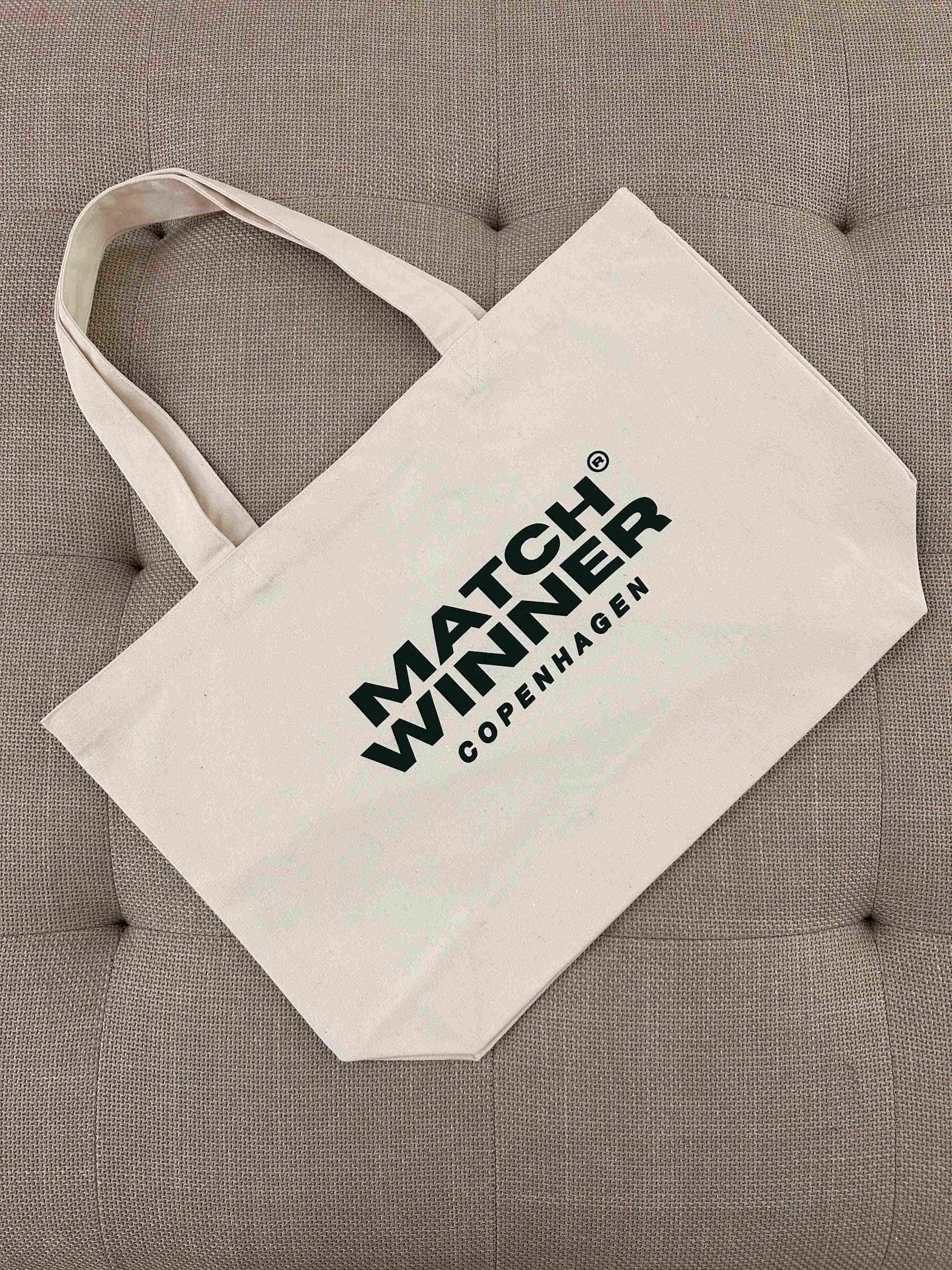 PREMIUM MW TOTEBAG fra Matchwinner i beige lærred med grønt logo liggende på en polstret bænk.